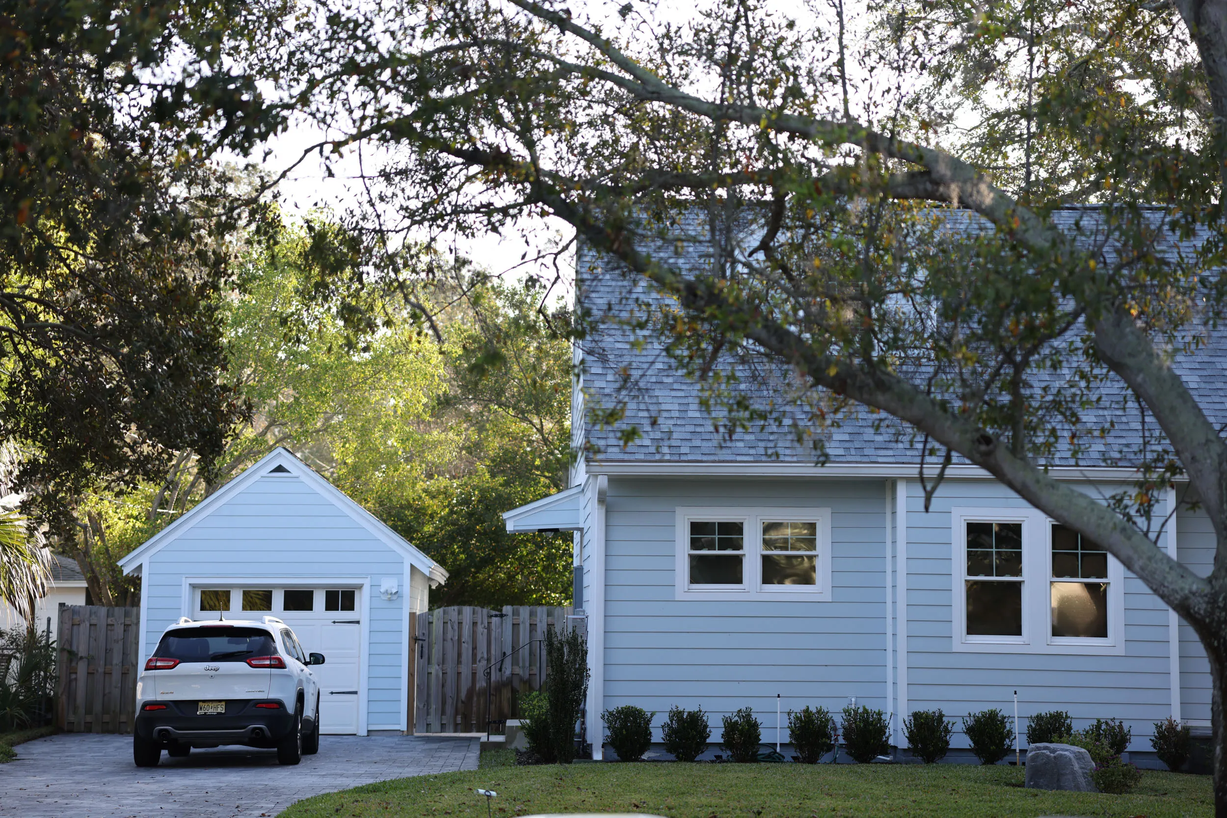 White Jeep SUV parked on driveway beside a light blue house and detached garage surrounded by trees.